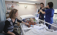 Mother sitting next to young boy in bed while two nurses attend
